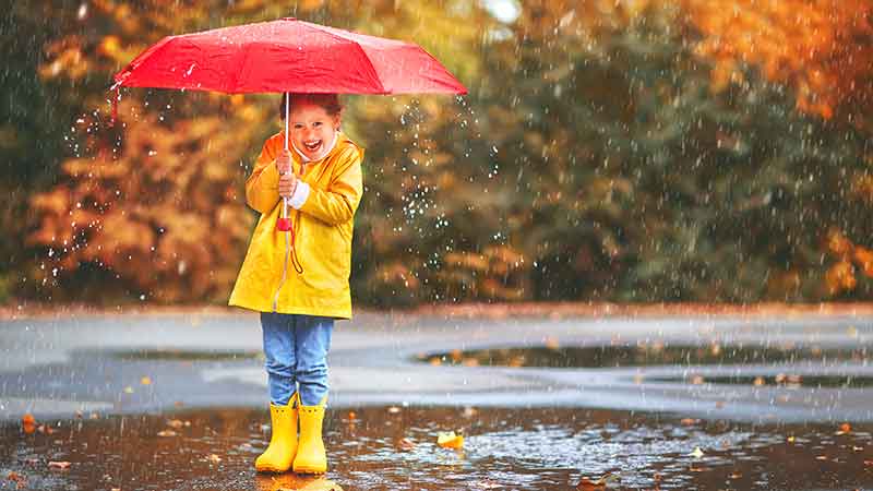 Child enjoying the rain with a red umbrella and yellow rain gear, highlighting the extensive protection of umbrella insurance.